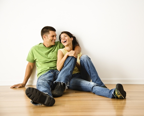 Attractive young adult couple sitting close on hardwood floor in home smiling and laughing.